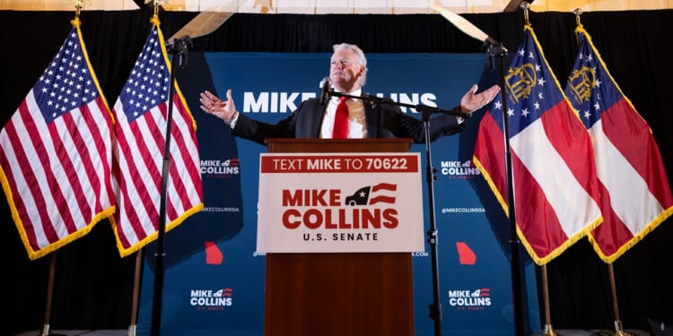 A man stands at a podium labeled Mike Collins U.S. Senate, arms outstretched, with American and Georgia state flags behind him and campaign signs in the background.