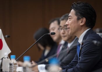 A man in a suit speaks at a conference table with other people, microphones, and a Japanese flag visible. Bottled water is on the table and everyone faces the same direction.
