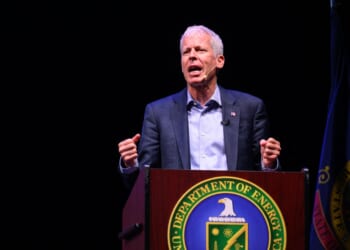 A man speaks passionately at a podium with the U.S. Department of Energy seal, flanked by the American flag and another flag, against a dark background.