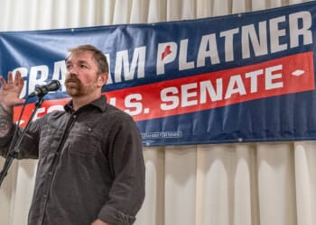 A man stands at a microphone with one hand raised, speaking in front of a campaign banner that reads Graham Platner U.S. Senate against a backdrop of white curtains.