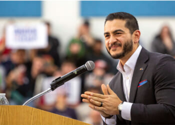 Abdul El-Sayed stands smiling at a podium in a dark suit.