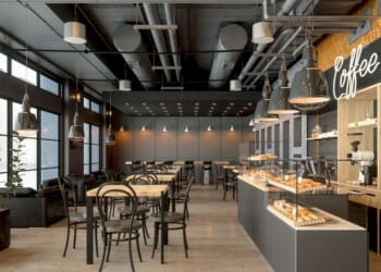 A coffee shop interior with wooden tables, a coffee maker, pastries, and pendant lights.