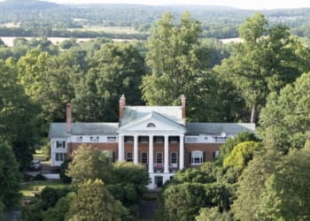 A large, historic mansion with white columns and red brick walls is surrounded by lush green trees, with rolling hills and fields visible in the background under a partly cloudy sky.