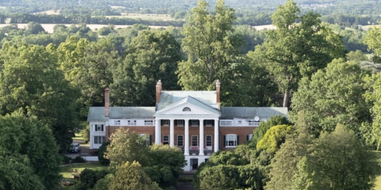 A large, historic mansion with white columns and red brick walls is surrounded by lush green trees, with rolling hills and fields visible in the background under a partly cloudy sky.