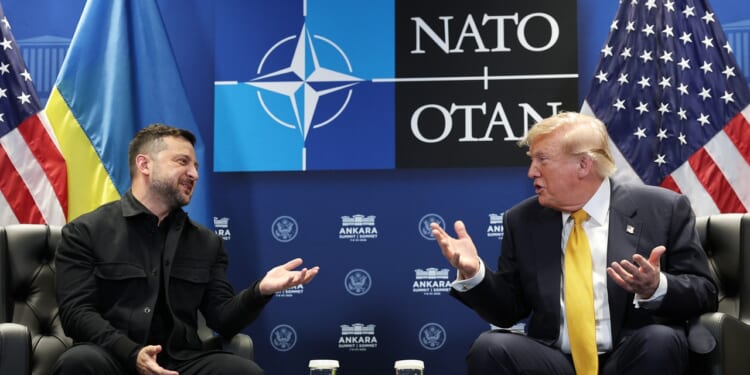 Two men in suits sit facing each other, gesturing while talking. Behind them are Ukrainian, NATO, and U.S. flags, along with a NATO OTAN logo.