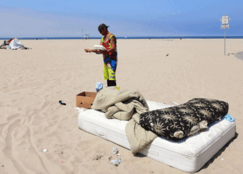 A person stands on a beach next to a pile of bedding and a box. There is a mattress, pillow, and blanket on the sand. Another person rides a bike in the background. The sky is clear and blue.