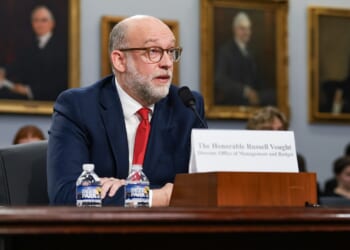 A man in a suit and red tie speaks into a microphone at a hearing, seated at a table with nameplate and water bottles. Portraits hang on the wall behind him.