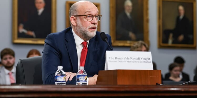 A man in a suit and red tie speaks into a microphone at a hearing, seated at a table with nameplate and water bottles. Portraits hang on the wall behind him.