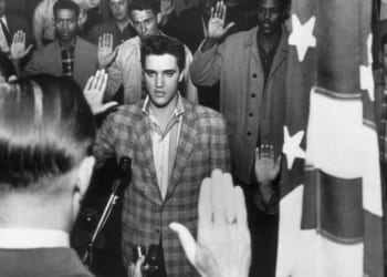 Elvis Presley stands with a group of young men at an induction center, raising their right hands as they are sworn into the United States Army by an officer standing next to an American flag.