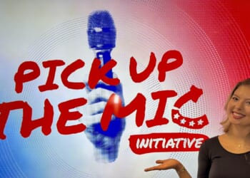 A woman smiles and gestures toward a screen displaying “PICK UP THE MIC INITIATIVE” in bold red letters with a graphic of a hand holding a microphone in the background.
