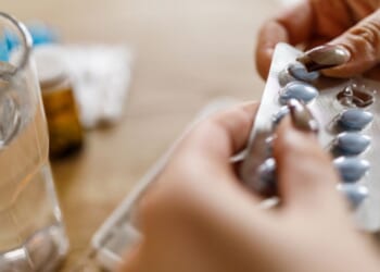 A woman opens a pill packet next to a cup of water sitting on a wooden table.
