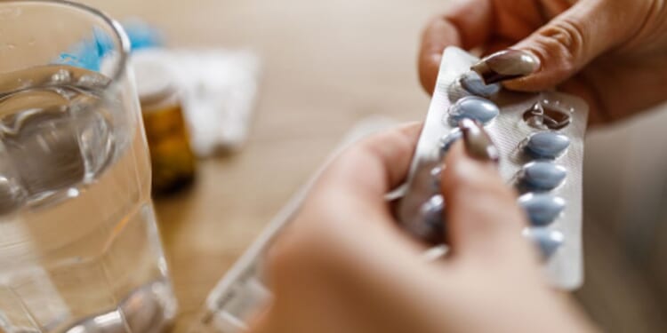 A woman opens a pill packet next to a cup of water sitting on a wooden table.
