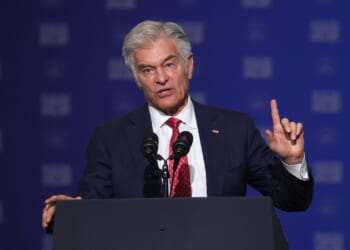 A man in a dark suit and red tie speaks at a podium with two microphones, gesturing with his right hand raised. The background is blue with white text.