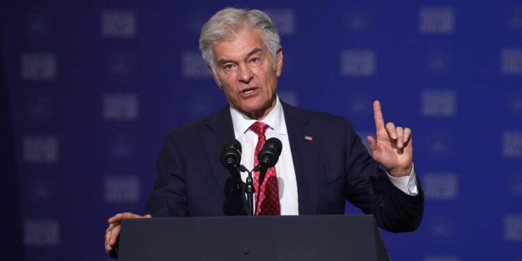 A man in a dark suit and red tie speaks at a podium with two microphones, gesturing with his right hand raised. The background is blue with white text.