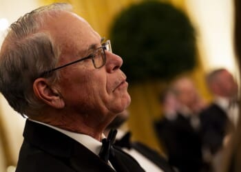 An older man wearing glasses and a tuxedo listens attentively at a formal event, with other people in tuxedos blurred in the background.