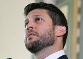 Florida Attorney General James Uthmeier is suing pro-transgender activist groups the American Academy of Pediatrics, the World Professional Association for Transgender Health, and the Endocrine Society. In this picture, Uthmeier with short dark hair and a beard, wearing a suit, looks upward while speaking indoors.