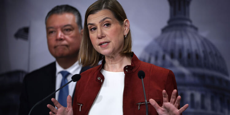 Sen. Elissa Slotkin says the SAVE Act would make it harder for "any Democrat in any state to win any election." A woman in a red jacket speaks at a podium with a man in a suit standing behind her. The blurred background includes a large government building dome and a U.S. flag.