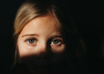 A close-up of a young child with blonde hair, partially illuminated by sunlight, with shadows obscuring the lower half of their face. The child looks directly at the camera with a serious, school-aged expression.