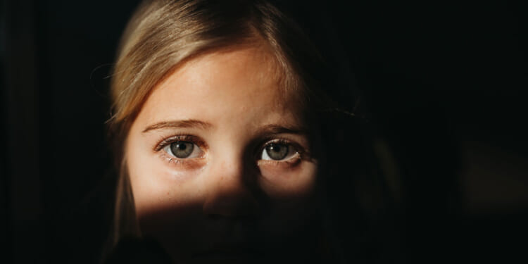 A close-up of a young child with blonde hair, partially illuminated by sunlight, with shadows obscuring the lower half of their face. The child looks directly at the camera with a serious, school-aged expression.
