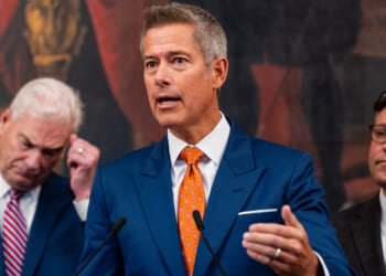 Department of Transportation Secretary Sean Duffy stands at a podium in a dark blue suit as he gestures with his hand. Rep. Tom Emmer and Speaker Mike Johnson stand behind him.