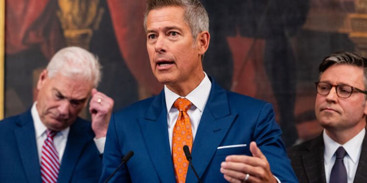 Department of Transportation Secretary Sean Duffy stands at a podium in a dark blue suit as he gestures with his hand. Rep. Tom Emmer and Speaker Mike Johnson stand behind him.