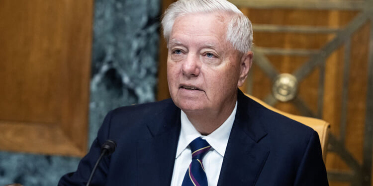 An older man in a suit and striped tie sits at a desk with a microphone, appearing to speak or listen during a formal meeting in a wood-paneled room.