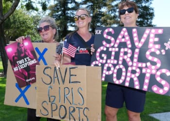 Three women stand outdoors holding signs reading SAVE GIRLS SPORTS and TWO XS NO EXCEPTIONS, showing support for girls sports. They smile and wear sunglasses, with trees and grass in the background.