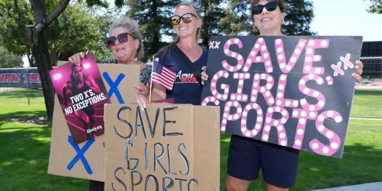 Three women stand outdoors holding signs reading SAVE GIRLS SPORTS and TWO XS NO EXCEPTIONS, showing support for girls sports. They smile and wear sunglasses, with trees and grass in the background.