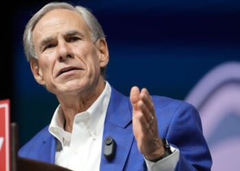 An older man in a blue checked jacket and white shirt speaks at a podium, gesturing with his right hand. A microphone is clipped to his jacket, and a sign is partially visible in the foreground.