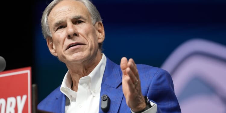 An older man in a blue checked jacket and white shirt speaks at a podium, gesturing with his right hand. A microphone is clipped to his jacket, and a sign is partially visible in the foreground.