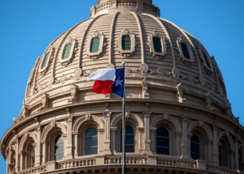 The Texas state flag waves in front of the ornate dome of the Texas State Capitol building under a clear blue sky.