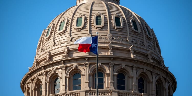 The Texas state flag waves in front of the ornate dome of the Texas State Capitol building under a clear blue sky.