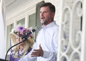 A man in a light blue shirt speaks at a microphone on a porch. Behind him, a woman in a decorative hat with flowers and a purple outfit listens. A white flag and part of a metal railing are visible.