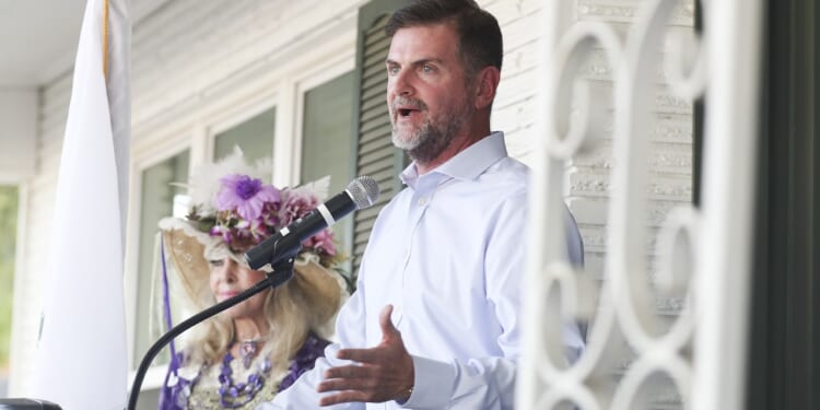 A man in a light blue shirt speaks at a microphone on a porch. Behind him, a woman in a decorative hat with flowers and a purple outfit listens. A white flag and part of a metal railing are visible.