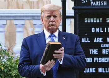 Trump holds up a closed book in front of a church sign outdoors. Shrubs and part of a building are visible in the background.
