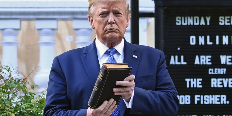 Trump holds up a closed book in front of a church sign outdoors. Shrubs and part of a building are visible in the background.