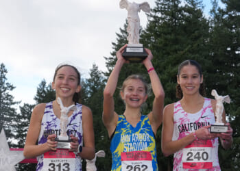 Three young female runners smile and hold white trophies, posing on a podium outdoors with trees in the background. All wear colorful running jerseys and race bibs—a vibrant testament to the spirit of Title IX, which opened doors for women in sports. The middle athlete raises her trophy above her head in celebration.