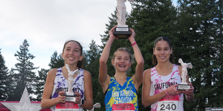 Three young female runners smile and hold white trophies, posing on a podium outdoors with trees in the background. All wear colorful running jerseys and race bibs—a vibrant testament to the spirit of Title IX, which opened doors for women in sports. The middle athlete raises her trophy above her head in celebration.
