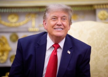 A man in a navy suit with a red tie sits on a light yellow patterned chair, smiling, in an ornate room with gold decorations.
