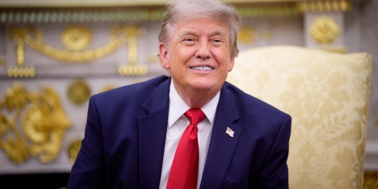 A man in a navy suit with a red tie sits on a light yellow patterned chair, smiling, in an ornate room with gold decorations.