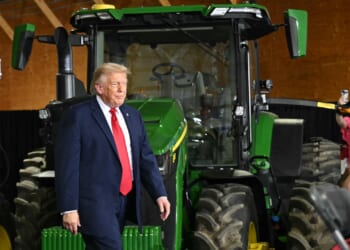 Trump walks past a large green tractor indoors, while people nearby watch and take photos.