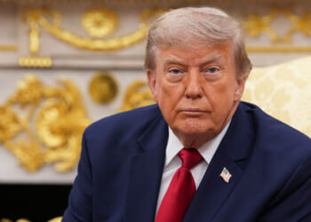 A man in a navy suit with a red tie and U.S. flag pin sits in an ornate room with gold decorations, looking forward with a serious expression.