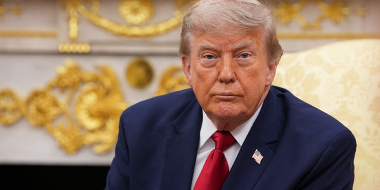 A man in a navy suit with a red tie and U.S. flag pin sits in an ornate room with gold decorations, looking forward with a serious expression.