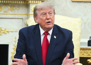 A man in a navy suit and red tie sits on a pale yellow chair, gesturing with his hands, in an ornate room with gold details and a dark bust in the background.