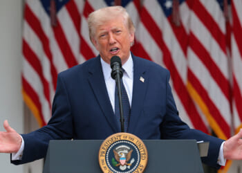 A man in a dark suit speaks at a podium with the U.S. presidential seal, gesturing with open arms. American flags are displayed in the background.