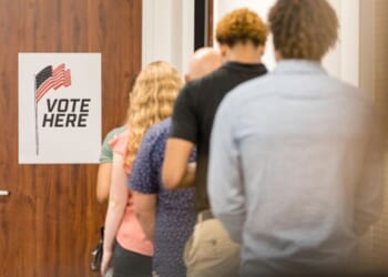 A line of multicultural citizens stand behind a door that has a sign that reads “vote here.”