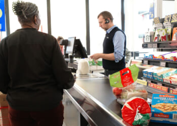 A cashier scans groceries, including bread and cheese, while a woman waits at the register in a brightly lit supermarket.