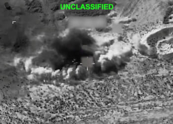 Black-and-white aerial view shows a large explosion with smoke billowing from the ground in a rugged, desert-like landscape. The word UNCLASSIFIED is displayed at the top in green text.