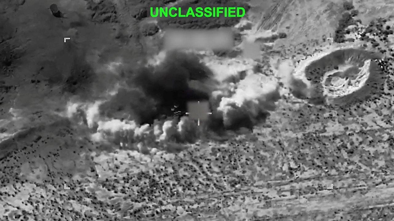 Black-and-white aerial view shows a large explosion with smoke billowing from the ground in a rugged, desert-like landscape. The word UNCLASSIFIED is displayed at the top in green text.