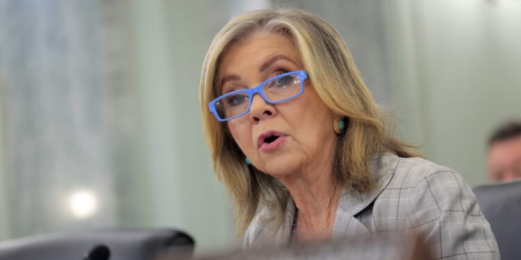A woman with shoulder-length blonde hair, blue glasses, and a plaid blazer speaks, appearing to be at a formal meeting or hearing.